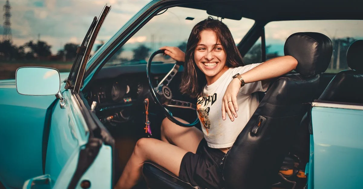 Une jeune femme joyeuse assise dans une voiture décapotable vintage bleue à l'extérieur pendant la journée.