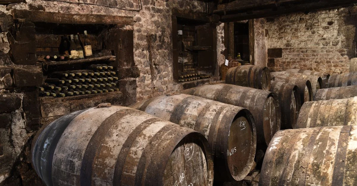 Une cave à vin atmosphérique avec des fûts en bois et des bouteilles poussiéreuses empilées contre des murs en pierre.